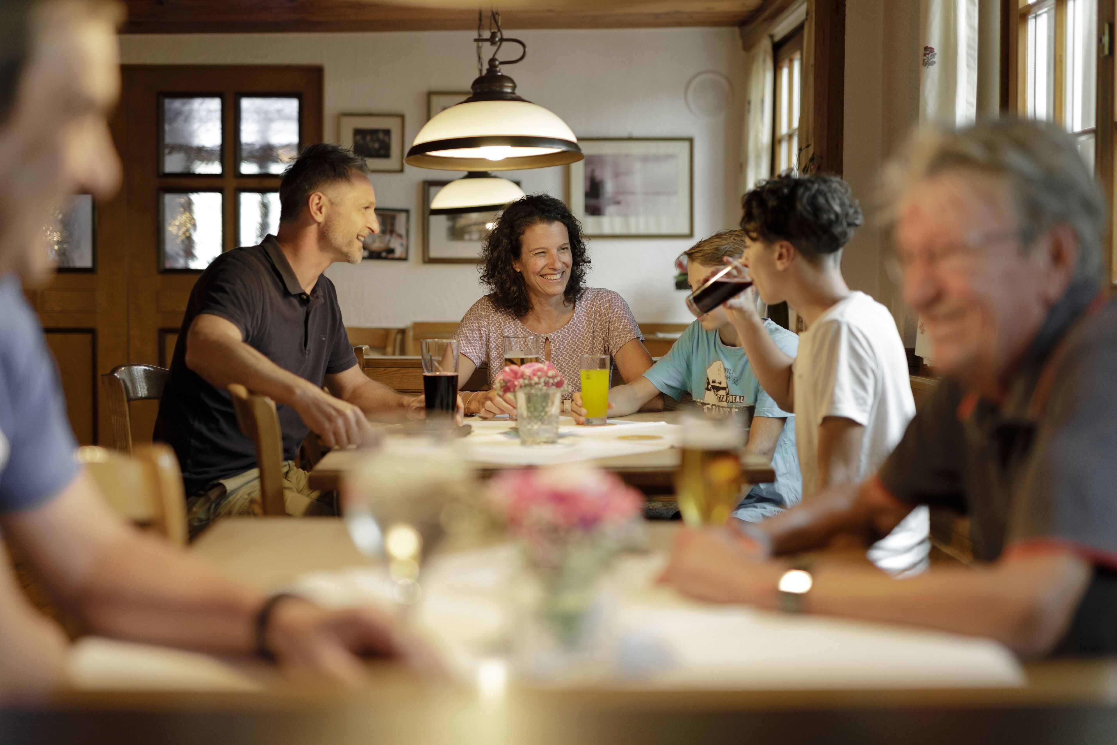 Un grupo de personas sentadas alrededor de una mesa en una habitación cálidamente iluminada, con muebles de madera y ventanas amplias.