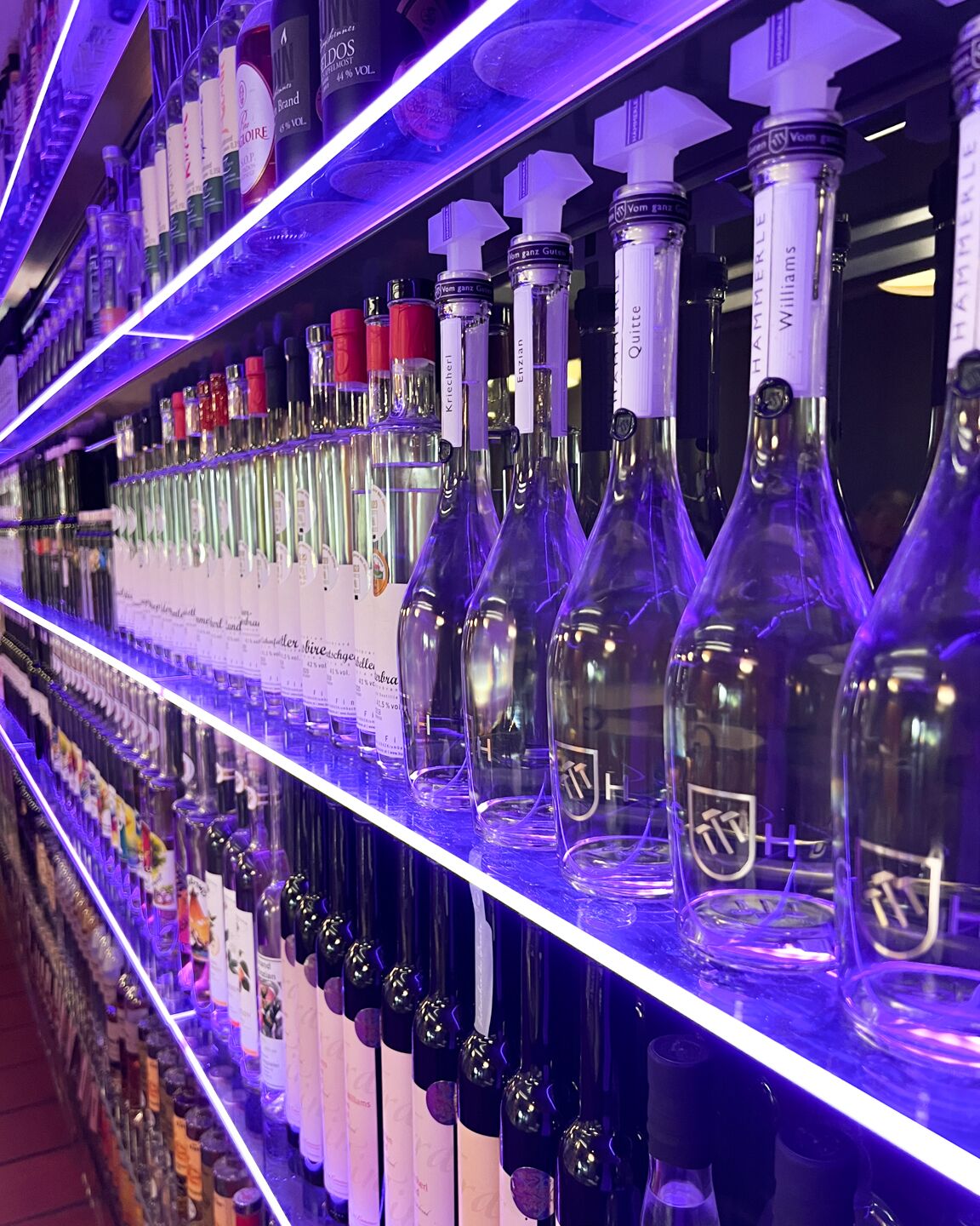 A bar shelf stocked with various liquor bottles, including wine and spirits, under purple lighting.