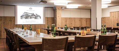 Un salón de conferencias con mesas largas y sillas, paredes de paneles de madera y una pantalla de proyector mostrando una imagen de hotel.