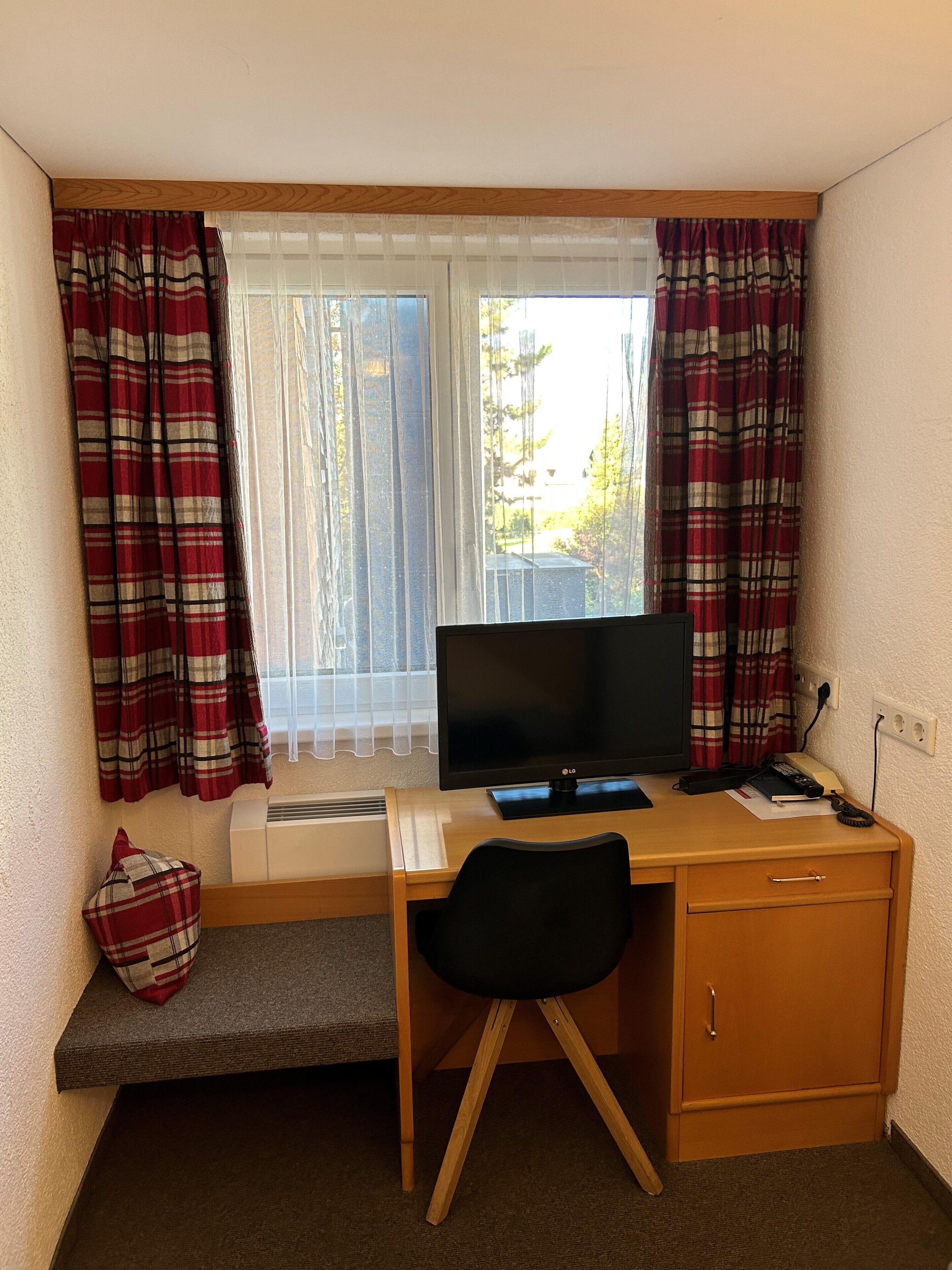 Un petit bureau comprenant un bureau, une chaise et un téléviseur. Il y a une fenêtre avec des rideaux, un radiateur et un petit banc.