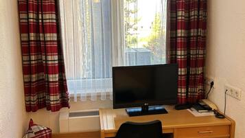 Ett litet kontorsutrymme med ett skrivbord, en stol och en tv. Det finns ett fönster med gardiner, ett element och en liten bänk.