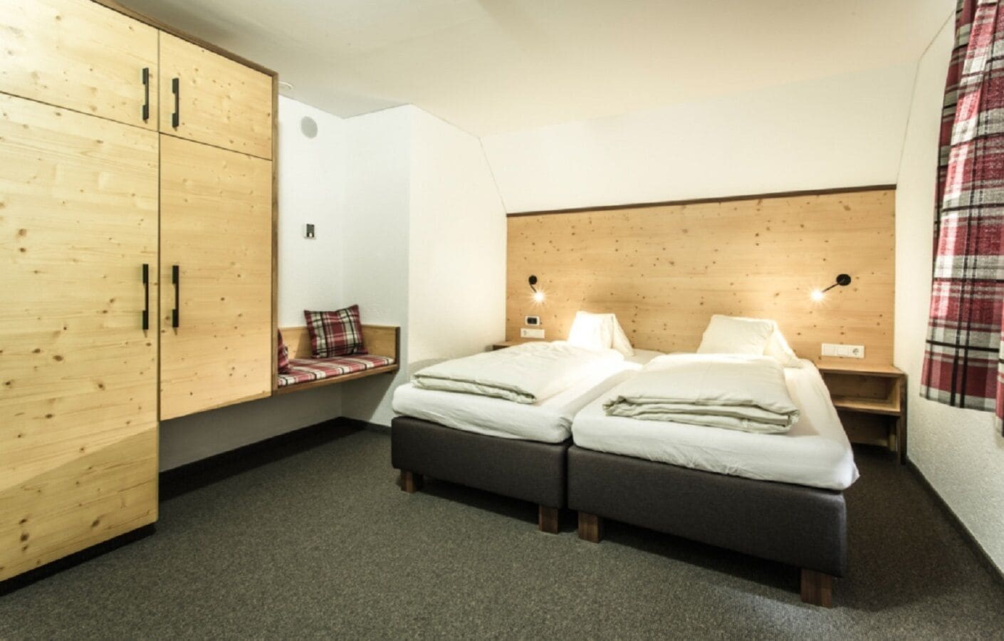 Una habitación con dos camas, un armario de madera, un banco con cojín escocés y una cortina a cuadros.