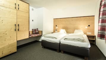 Una habitación con dos camas, un armario de madera, un banco con cojín escocés y una cortina a cuadros.