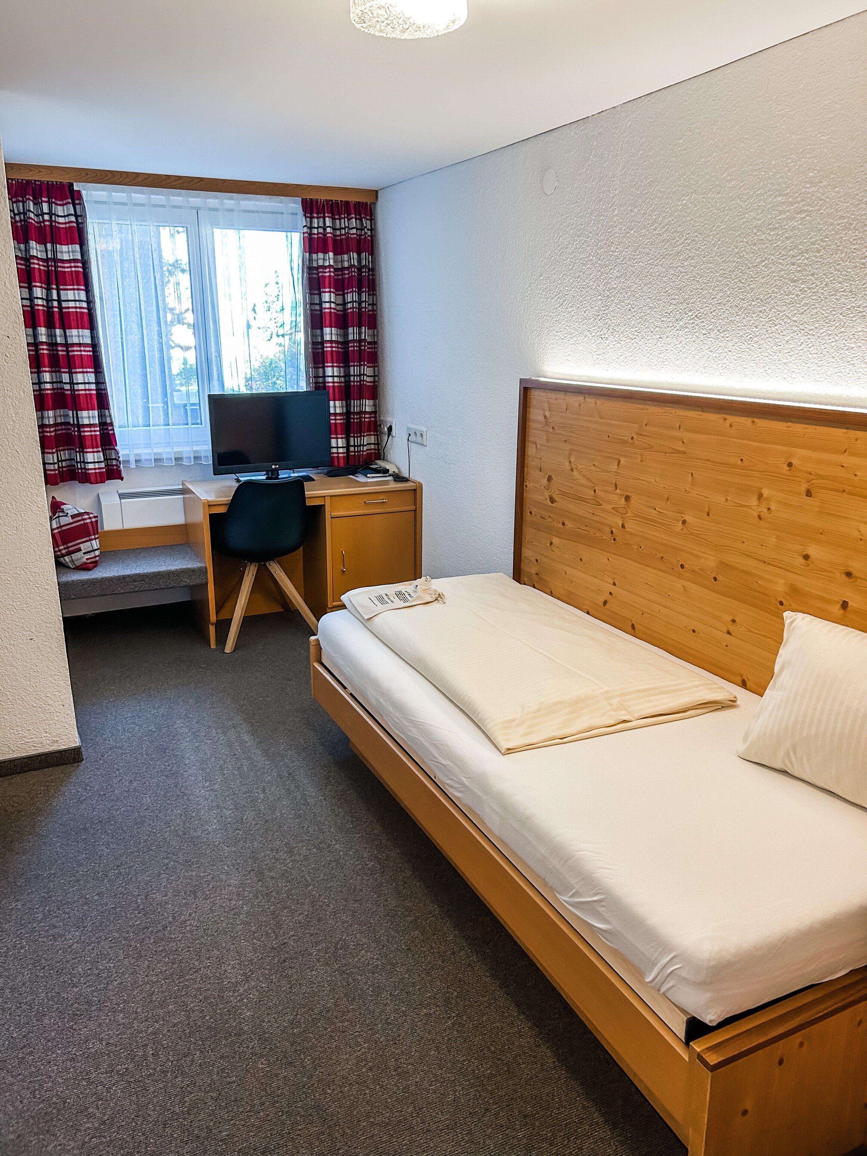 Ett hotellrum med en säng i trä, ett skrivbord med en dator, ett fönster med gardiner och en liten värmare.