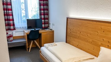 Ett hotellrum med en säng i trä, ett skrivbord med en dator, ett fönster med gardiner och en liten värmare.