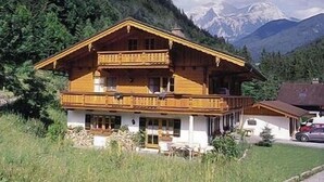 Un chalet in legno a due piani con un balcone, immerso nel verde e con montagne sullo sfondo.