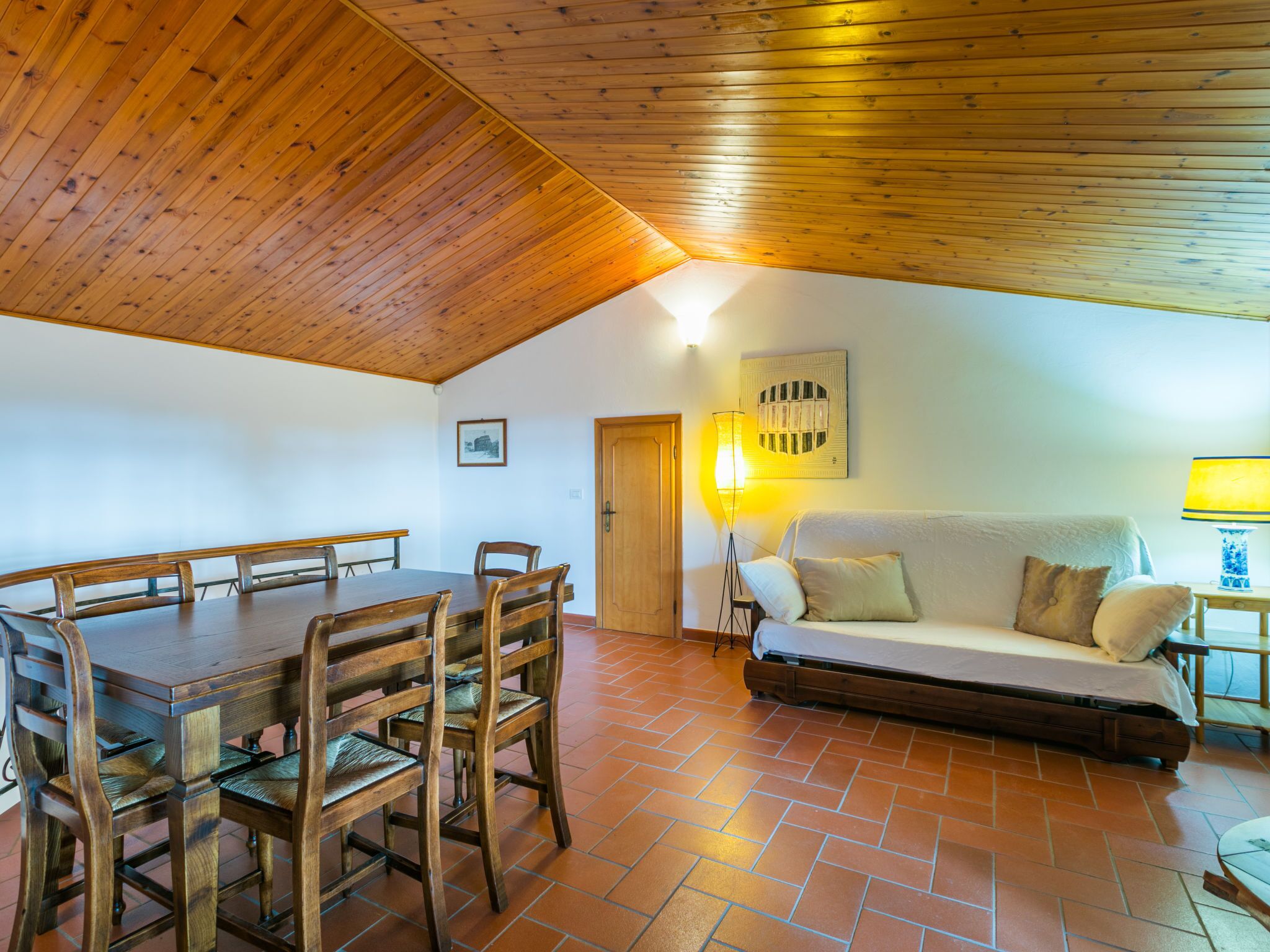 A cozy room with a wooden ceiling, a dining table with chairs, a sofa, and a lamp.