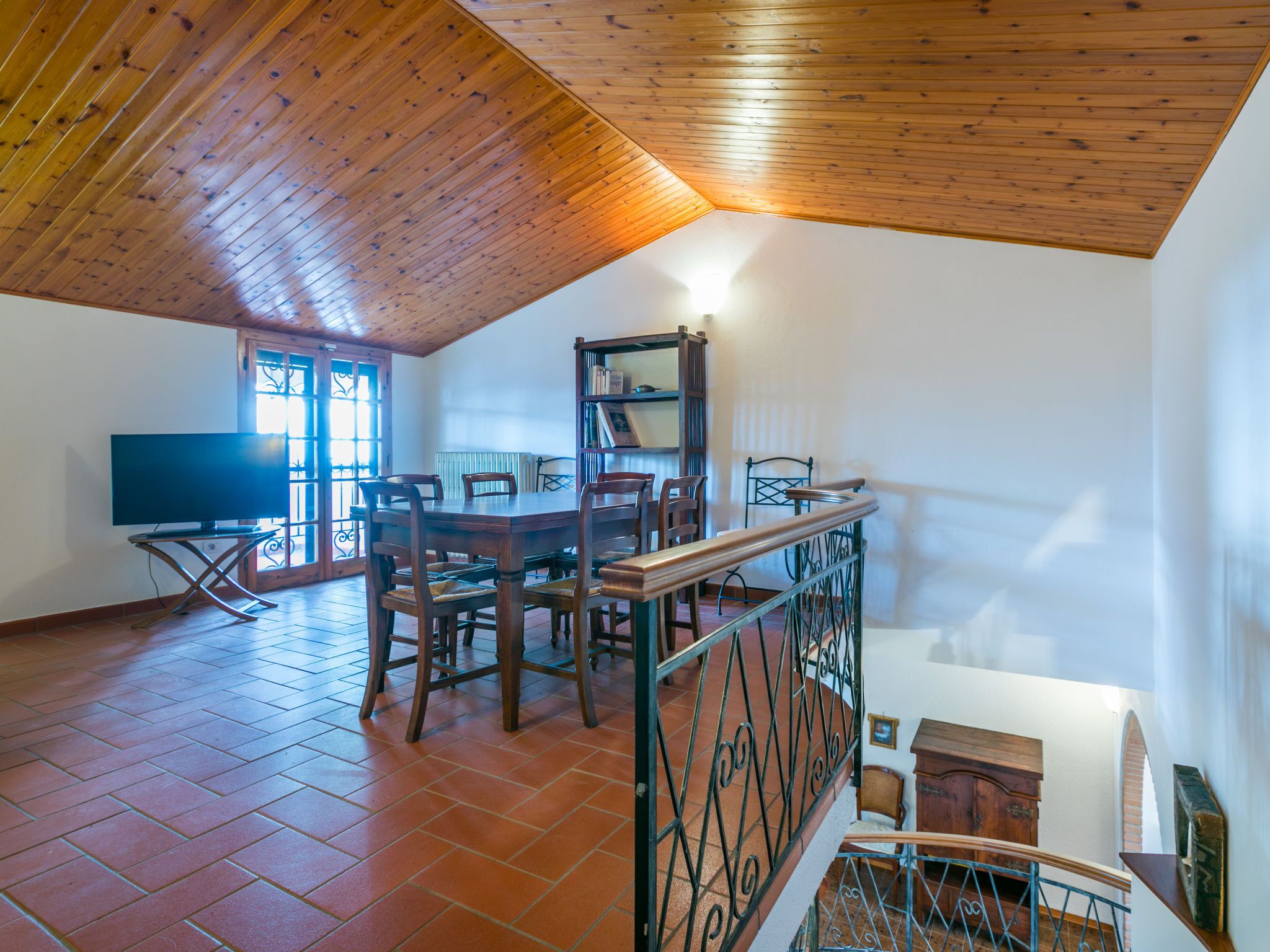 A spacious room with a wooden ceiling, a dining table with chairs, a television, and a bookshelf.