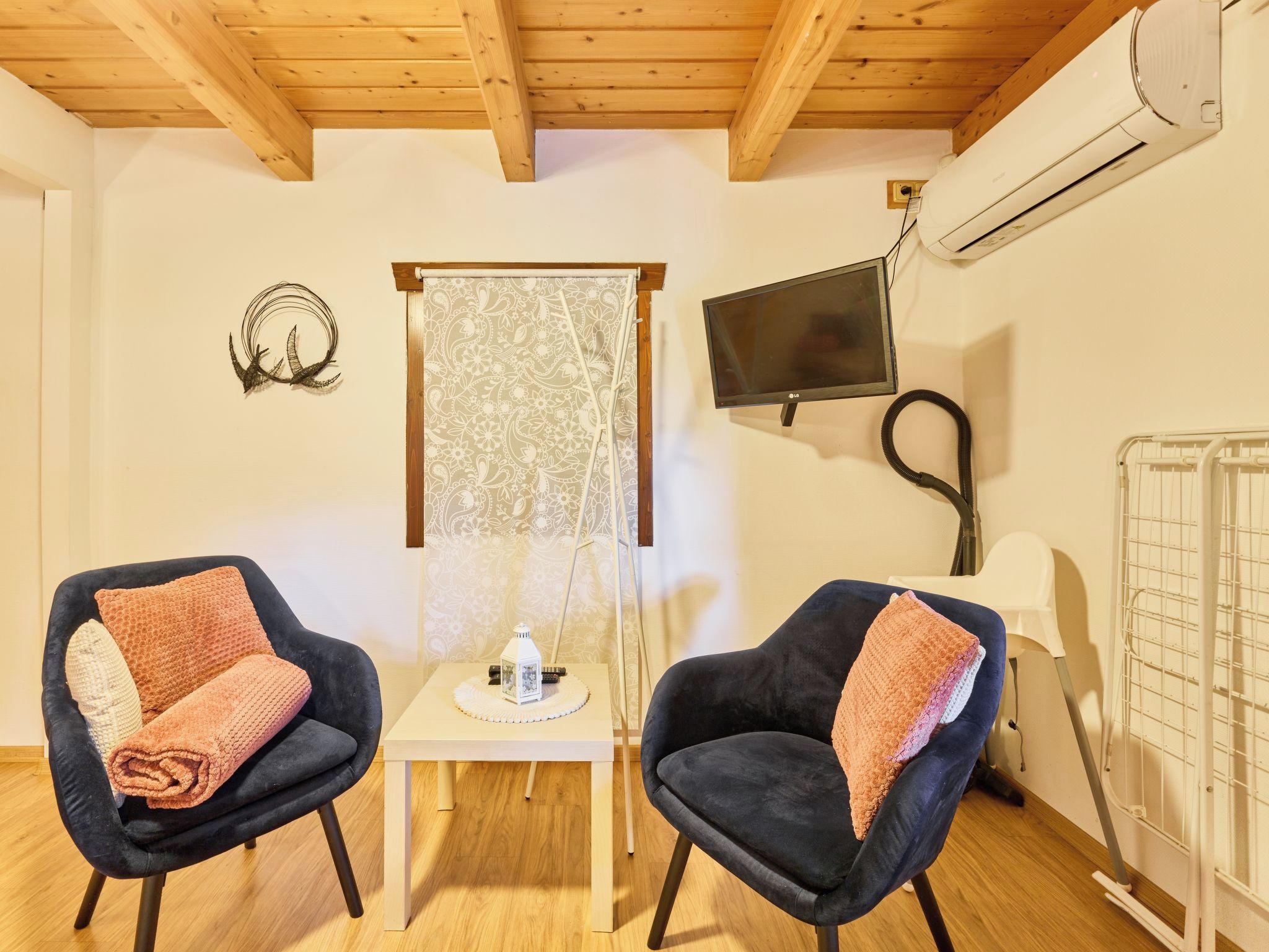 Una habitación con dos sillones oscuros, una mesita con un objeto decorativo, un televisor en la pared y un equipo de aire acondicionado.