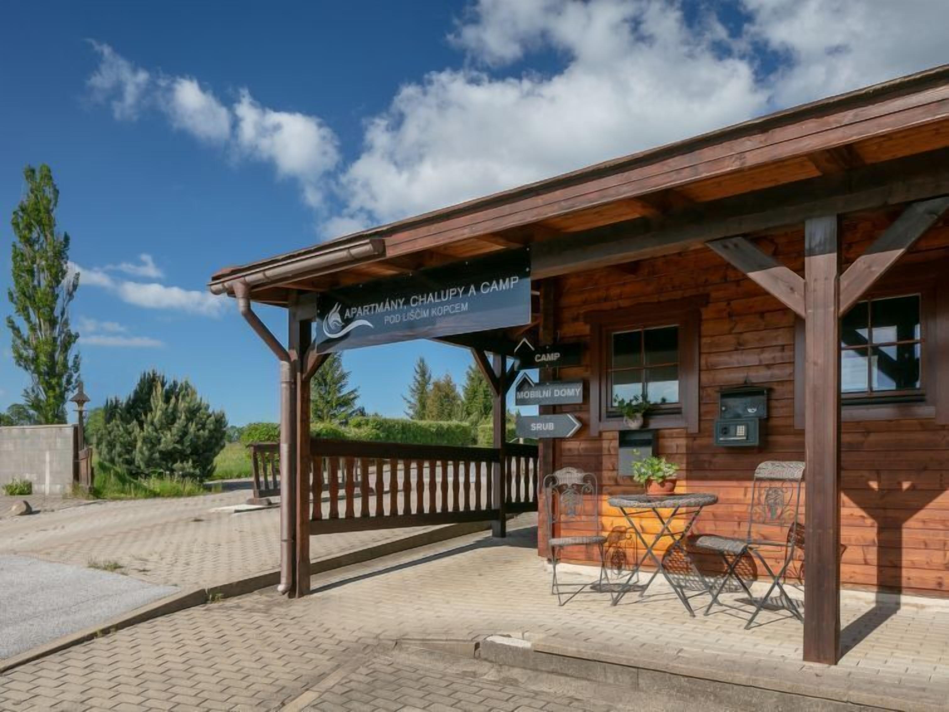 A wooden cabin with a sign indicating 'Apartment, Chalupy A Camp Pod Licem Kopcem'. There are outdoor tables and chairs, and a sign pointing to 'Camp' and 'Mobili Domy'.