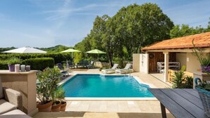 Un área de piscina con sillones de descanso y sombrillas, rodeada de vegetación y un edificio con techo de tejas.