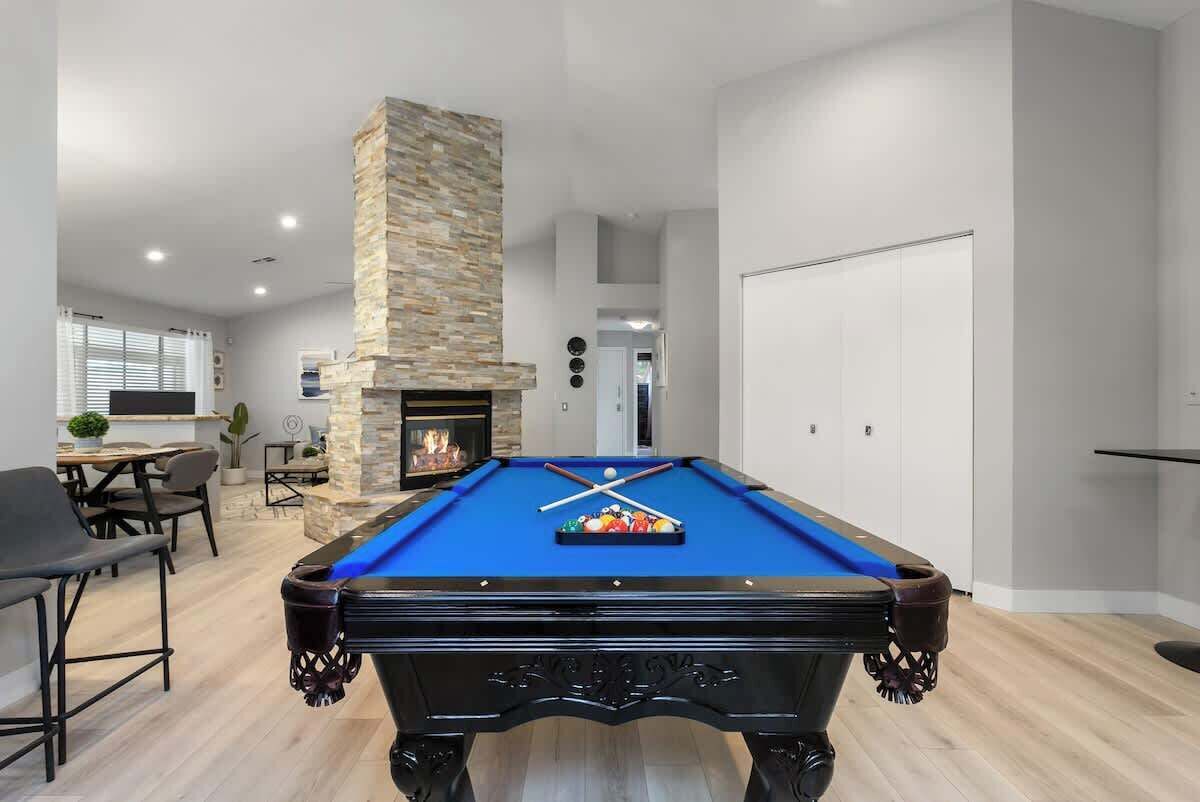 A pool table in a modern living room with a stone fireplace, a dining area, and a hallway leading to a door.