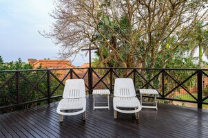 A wooden deck with two white lounge chairs and a small table, surrounded by a metal railing and lush greenery.
