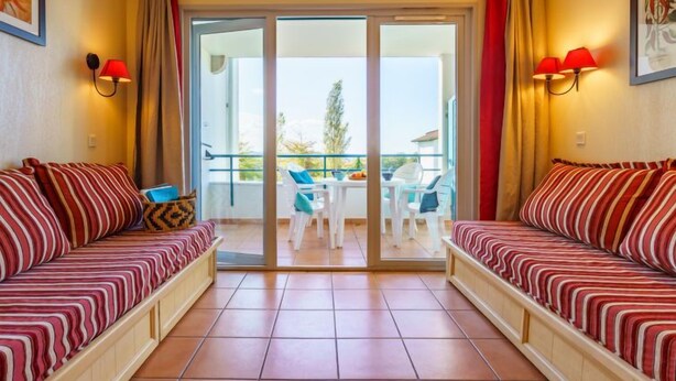 A hotel room with two striped sofas, a balcony with a table and chairs, and a large sliding glass door leading to the balcony. - Les Terrasses d'Arcangues by Interhome (Arcangues)