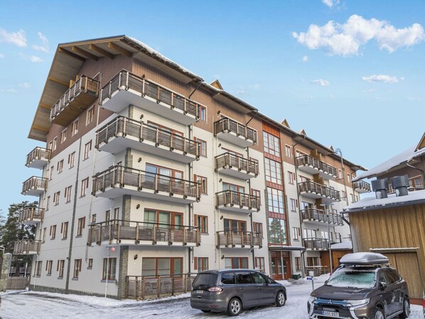 A multi-story building with balconies, a car parked in front, and snow on the ground.