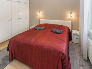 A neatly made bed with a red bedspread and two folded towels, a white headboard, bedside tables with lamps, and a wardrobe with sliding doors.