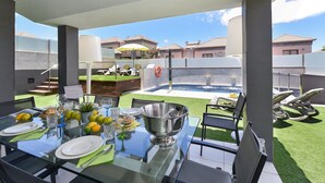 An outdoor dining area with a glass table, chairs, and a view of a swimming pool and buildings in the background. - Meloneras Hills 19 by Interhome (Maspalomas)