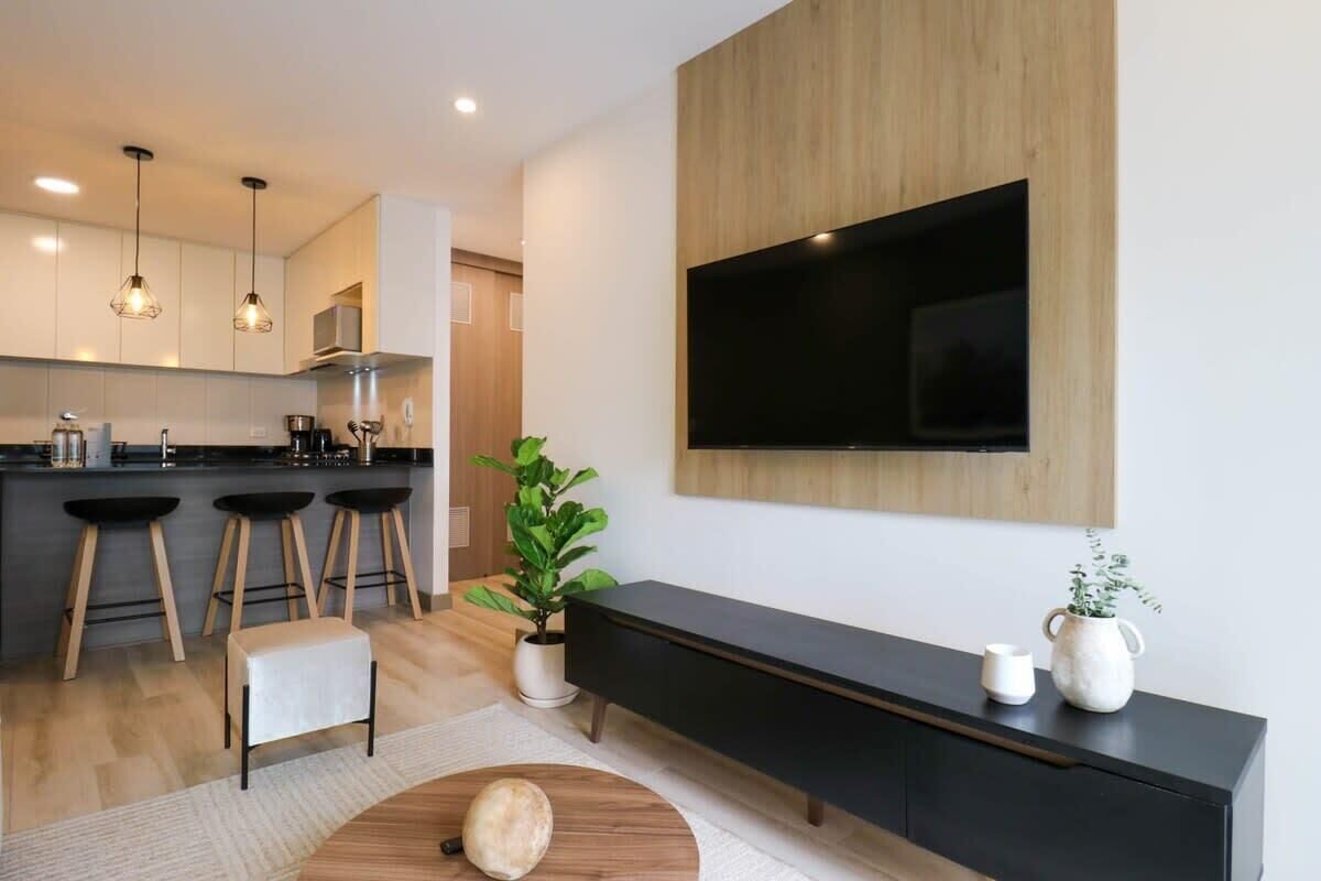 Une cuisine moderne avec des meubles en bois, un plan de travail noir et trois tabourets de bar. Un grand téléviseur à écran plat est fixé au mur, au-dessus d’une console noire.