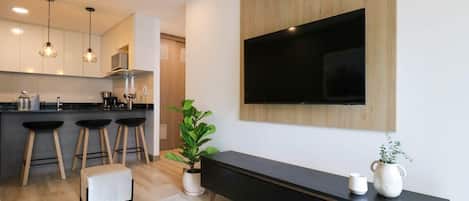 Una cocina moderna con armarios de madera, una encimera negra y tres taburetes de bar. Hay un televisor de pantalla plana grande montado en la pared sobre un mueble consola negro.