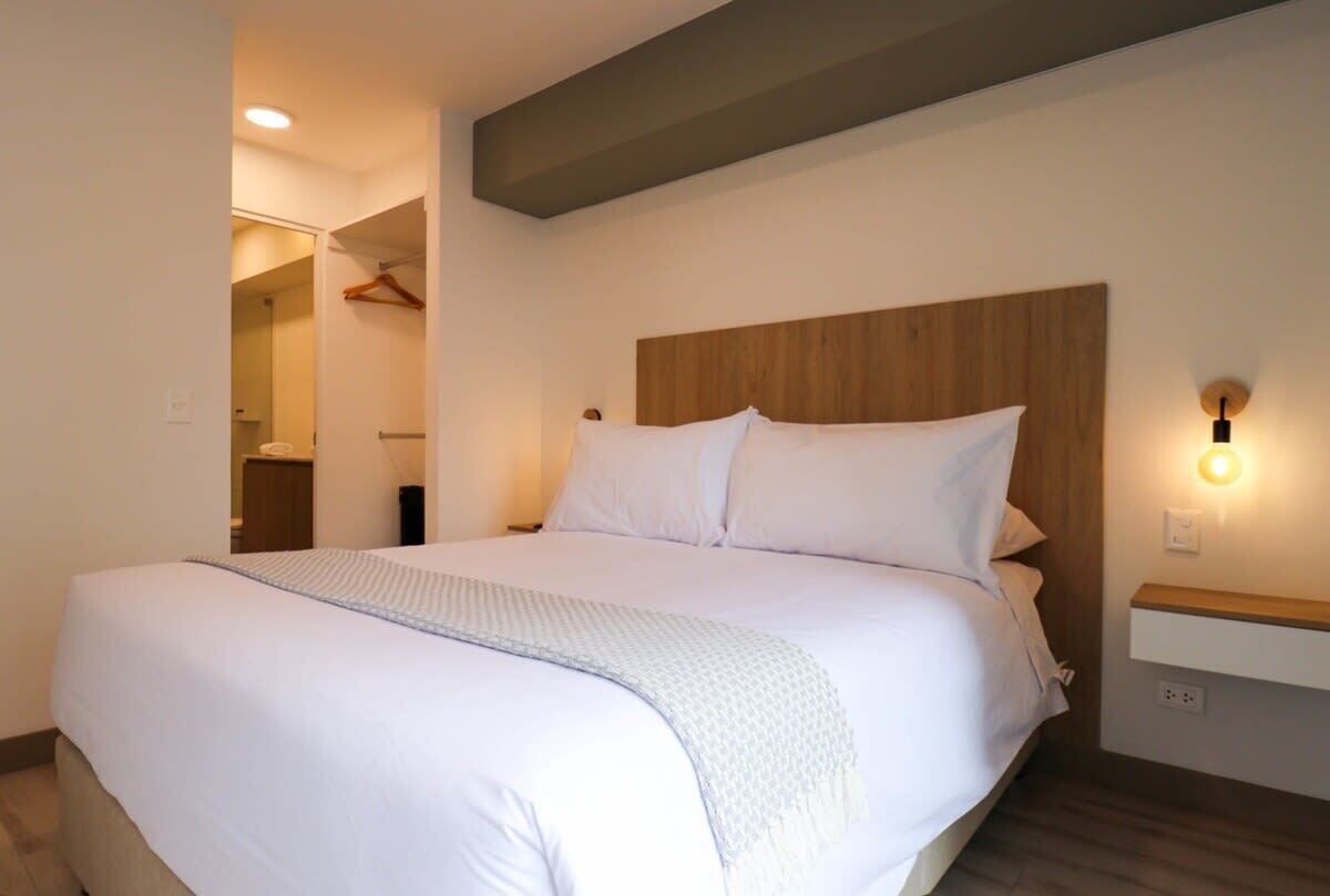 Una cama bien hecha con cabecera de madera, una mesita de noche con lámpara y un aplique de pared.