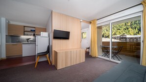 A compact room with a kitchenette, a flat-screen TV mounted on a wooden partition, a dining area with a table and chairs, and a sliding glass door leading to an outdoor deck with a view of a forested area. - Apt 4/6 ppl near the ski runs, mountain view (Allos)