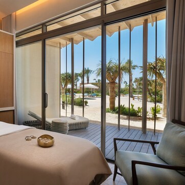 A modern hotel room with a bed, a large mirror, a bathroom with a sink, and a balcony with a view of palm trees and a beach.