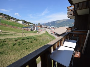 Une vue depuis un balcon donnant sur un village, avec des bâtiments, une zone herbeuse et des montagnes en arrière-plan.
