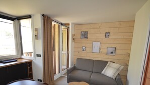 A compact living space with a sofa, a small wooden table, a bench, and a desk. There are framed pictures on the wall, a window with curtains, and a door leading to another room. - Renovated studio at the foot of the slopes with balcony and equipped kitchen (La Plagne Montalbert)