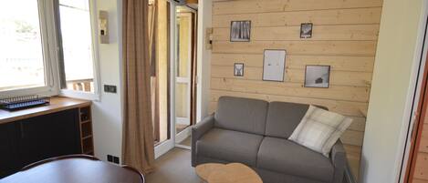 Un espacio habitable compacto con un sofá, una pequeña mesa de madera, un banco y un escritorio. Hay cuadros enmarcados en la pared, una ventana con cortinas y una puerta que conduce a otra habitación.