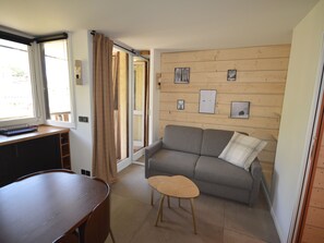 A compact living space with a sofa, a small wooden table, a bench, and a desk. There are framed pictures on the wall, a window with curtains, and a door leading to another room.