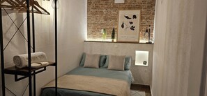 Ein kleines, gemütliches Schlafzimmer mit einem Einzelbett, einem Holzregal mit Handtüchern, einem gerahmten Pflanzenprint an der Wand und einer Backsteinwand.