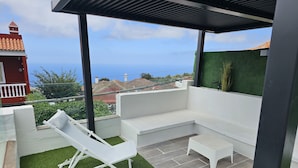 A covered outdoor seating area with a white sofa and lounge chairs, a small table, and a view of the sea.
