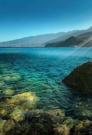 Eine Küstenansicht mit klarem Wasser, felsigem Ufer und Bergen im Hintergrund.