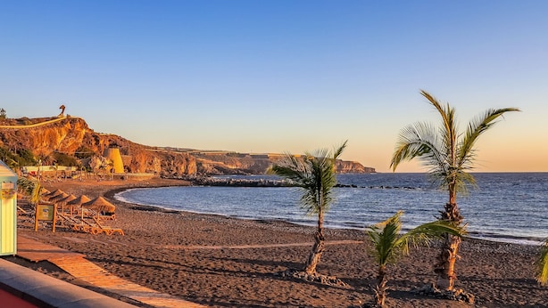 A beach with a rocky cliff, palm trees, and a wooden walkway leading to beach chairs.