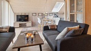 Een moderne woonkamer met een grijze bank, een houten salontafel en een televisie. Er is een eethoek met een tafel en stoelen, en een keuken op de achtergrond.