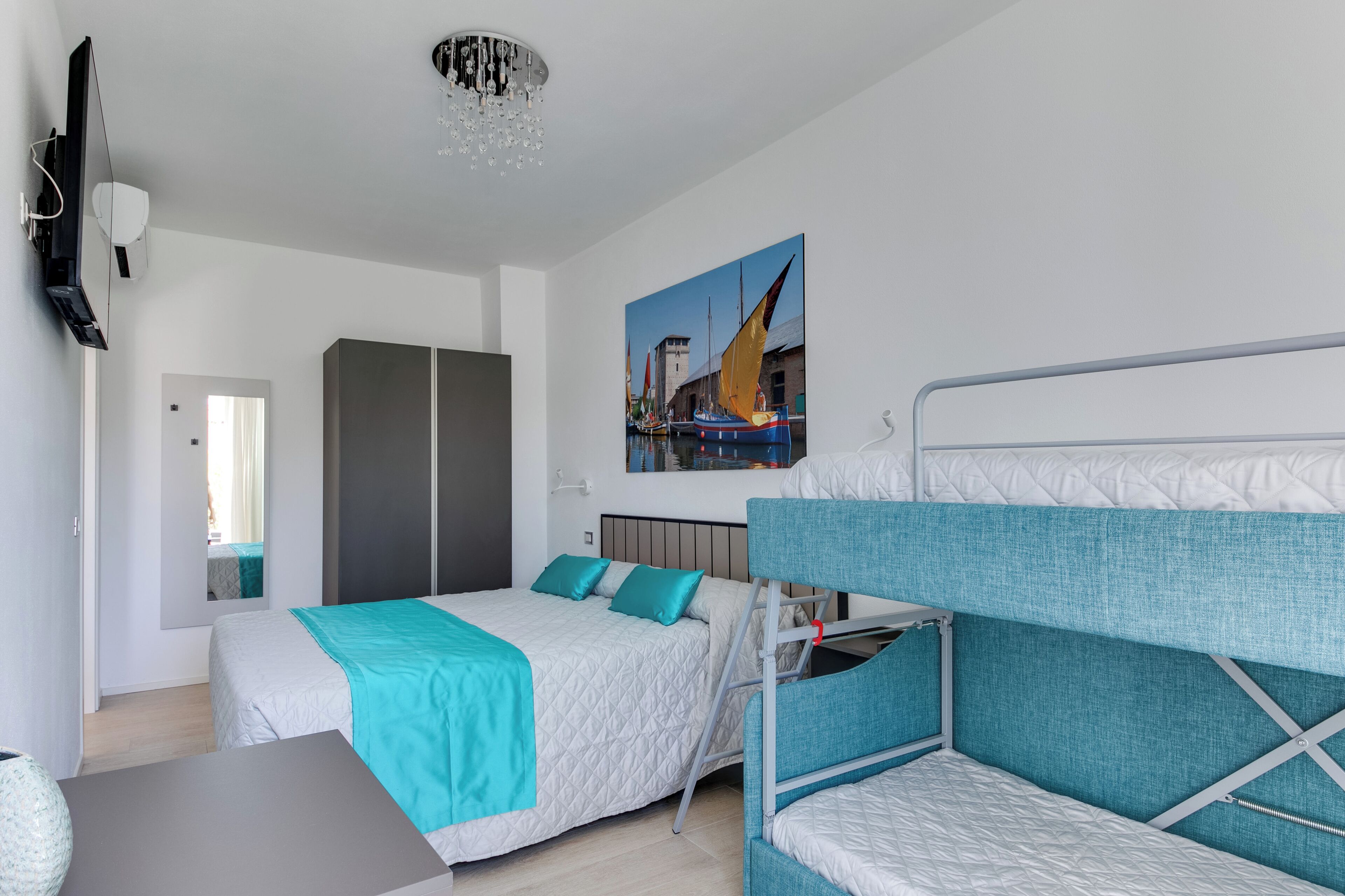 A modern hotel room with a bunk bed, a desk, a wardrobe, and a painting of boats on the wall.