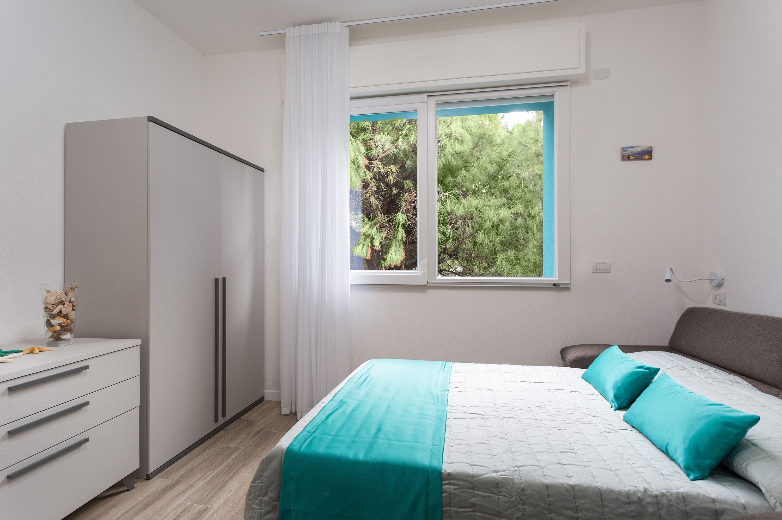Un dormitorio con cama, armario, ventana con vistas a árboles y una mesita con un florero.