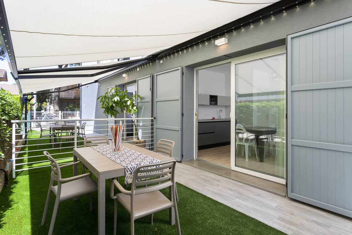 A covered outdoor dining area with a table and chairs, a potted plant, and a view into the house.