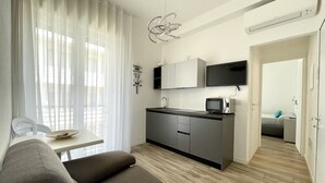 Un espacio habitable compacto con kitchenette, un sofá, una mesa de comedor y vistas a través de una ventana con cortinas traslúcidas.