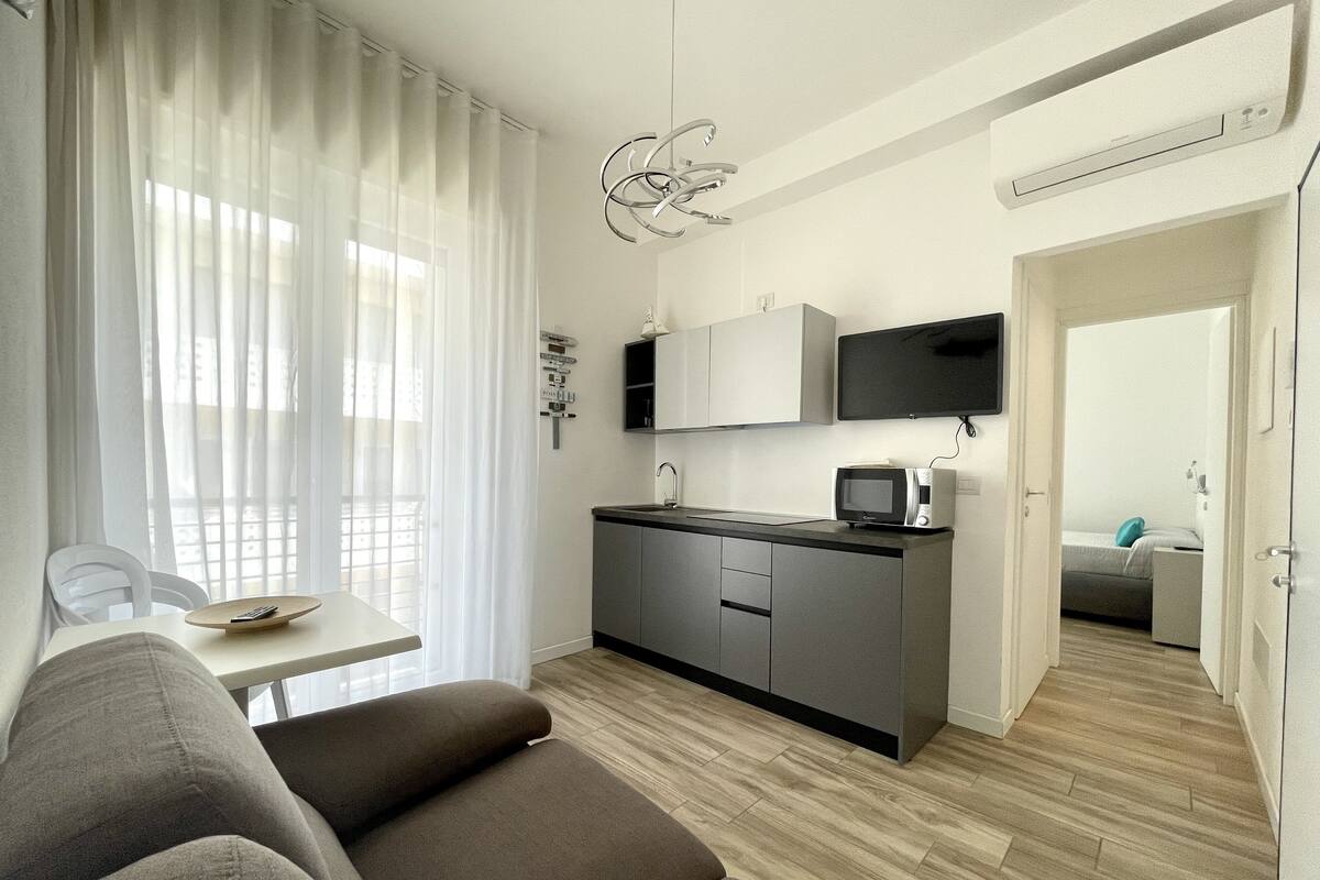 A compact living space with a kitchenette, a sofa, a dining table, and a view through a window with sheer curtains.