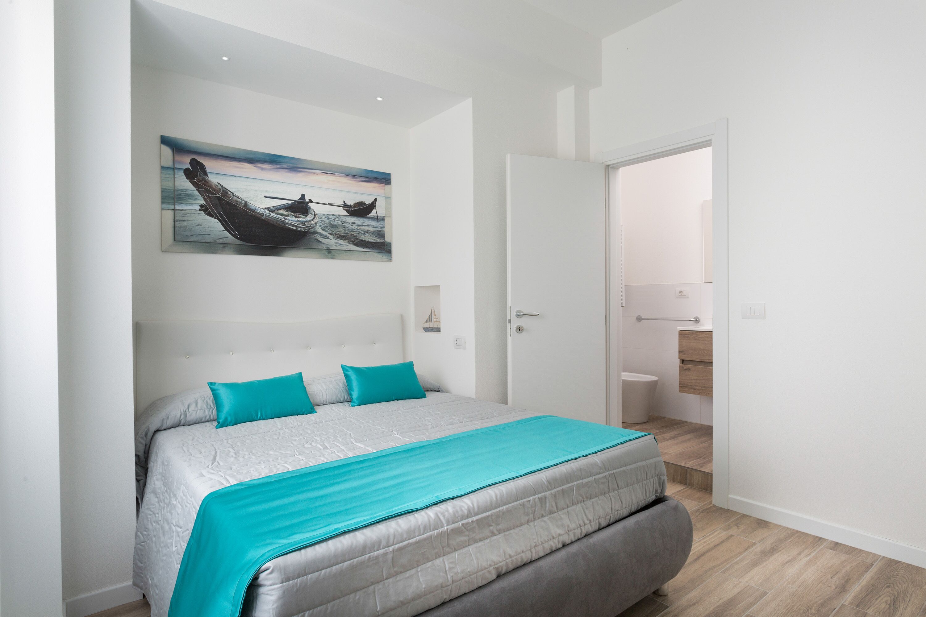 A modern bedroom with a large bed, a painting of a boat on the wall, and a view into the bathroom.
