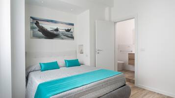 A modern bedroom with a large bed, a painting of a boat on the wall, and a view into the bathroom.