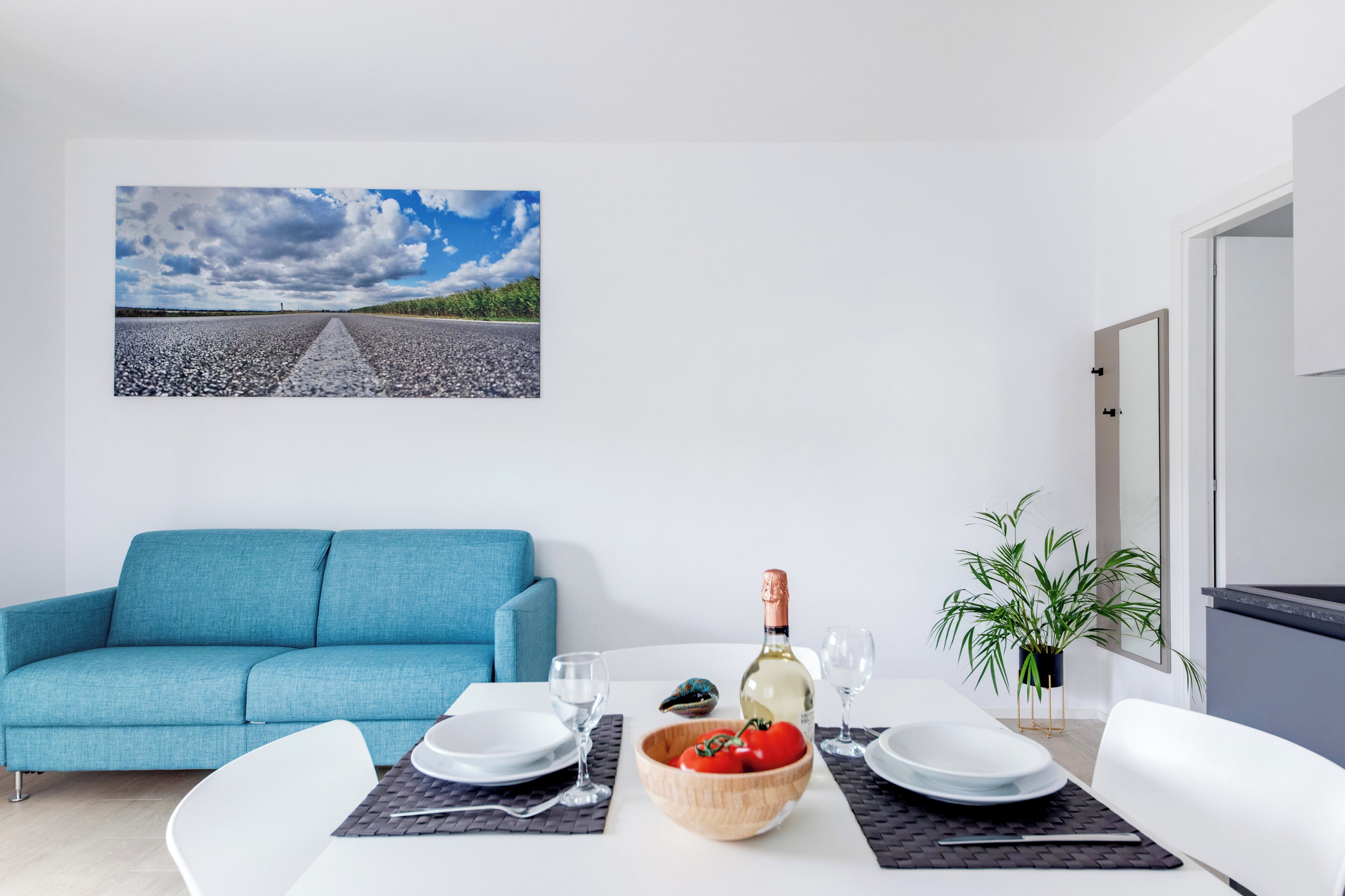Un soggiorno moderno con un divano blu, un tavolo da pranzo apparecchiato con piatti, bicchieri e una bottiglia di vino, e un'ampia finestra con vista su una strada e il cielo.