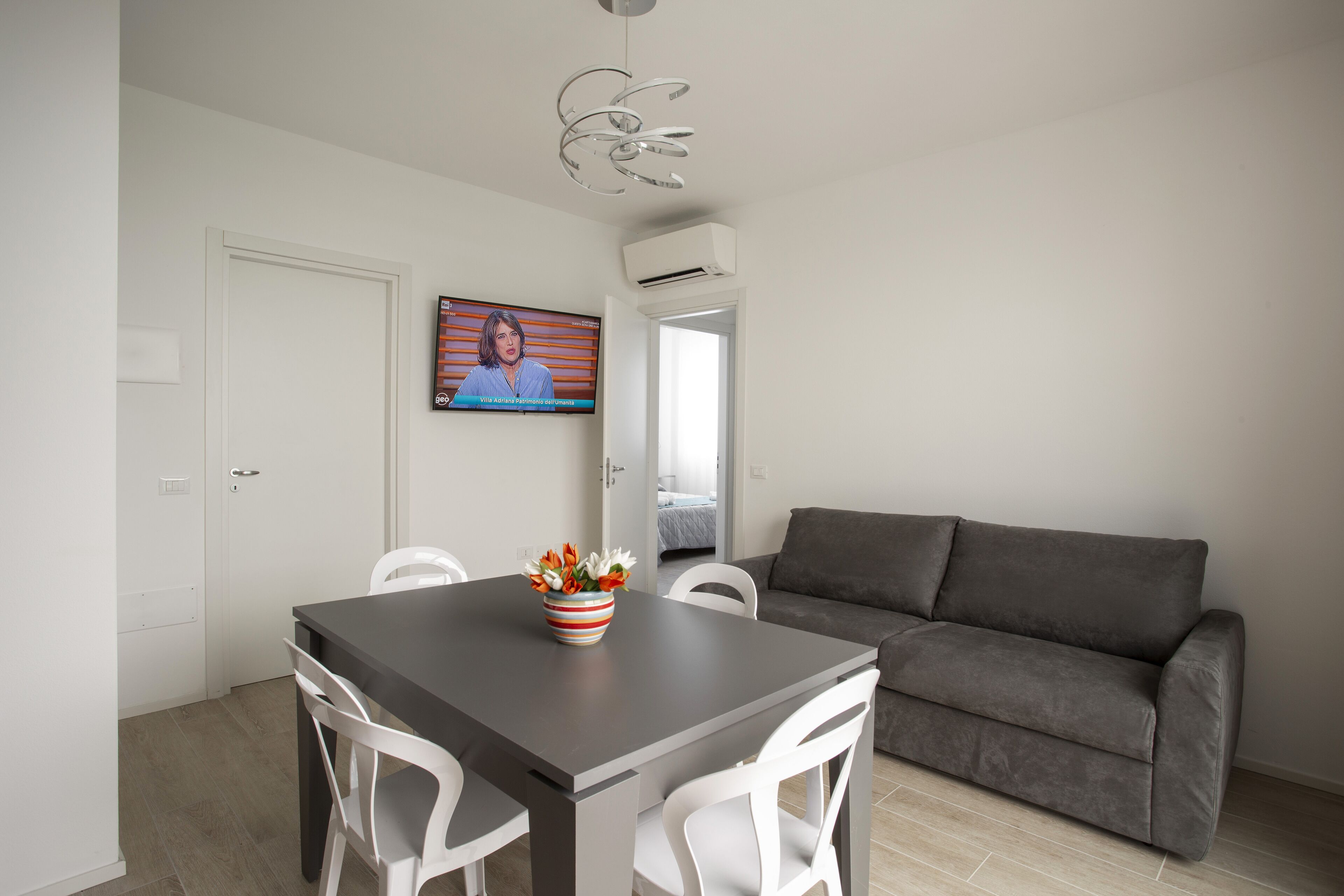 A modern living room with a grey sofa, white chairs, a dark wooden table with a striped vase, and a TV on the wall.