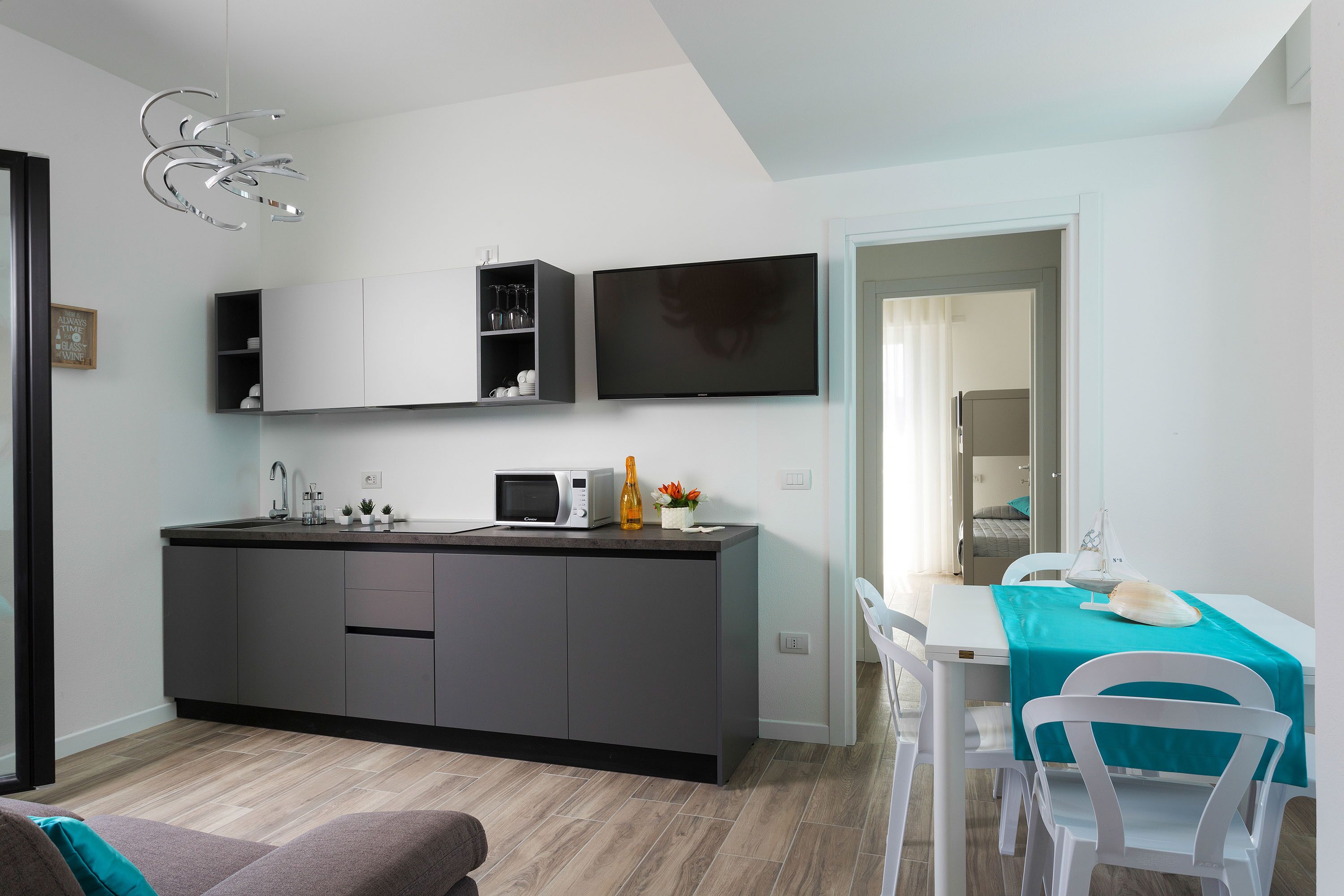 A modern kitchen with a built-in microwave, a sink, and a small cabinet. There is a dining area with a table and chairs, and a TV mounted on the wall.