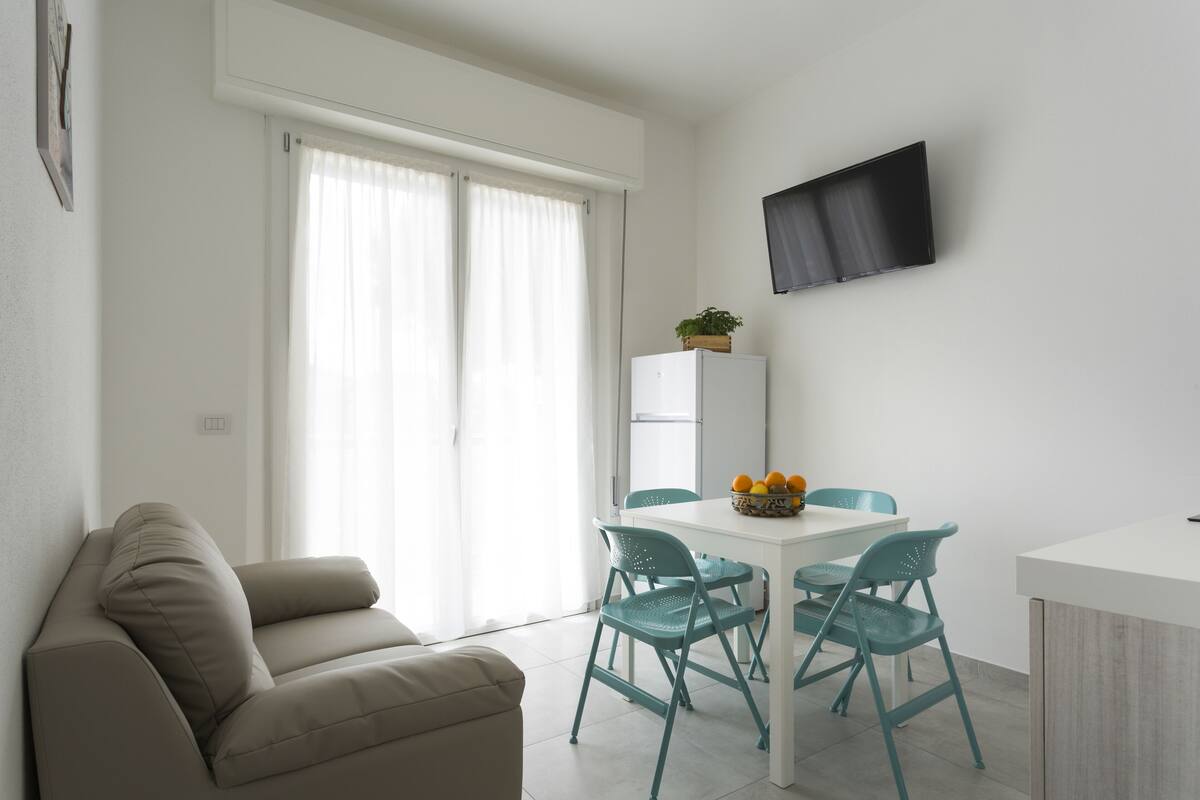 A modern living room with a sofa, a dining table with chairs, a flat-screen TV mounted on the wall, and a refrigerator.