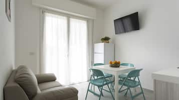A modern living room with a sofa, a dining table with chairs, a flat-screen TV mounted on the wall, and a refrigerator.