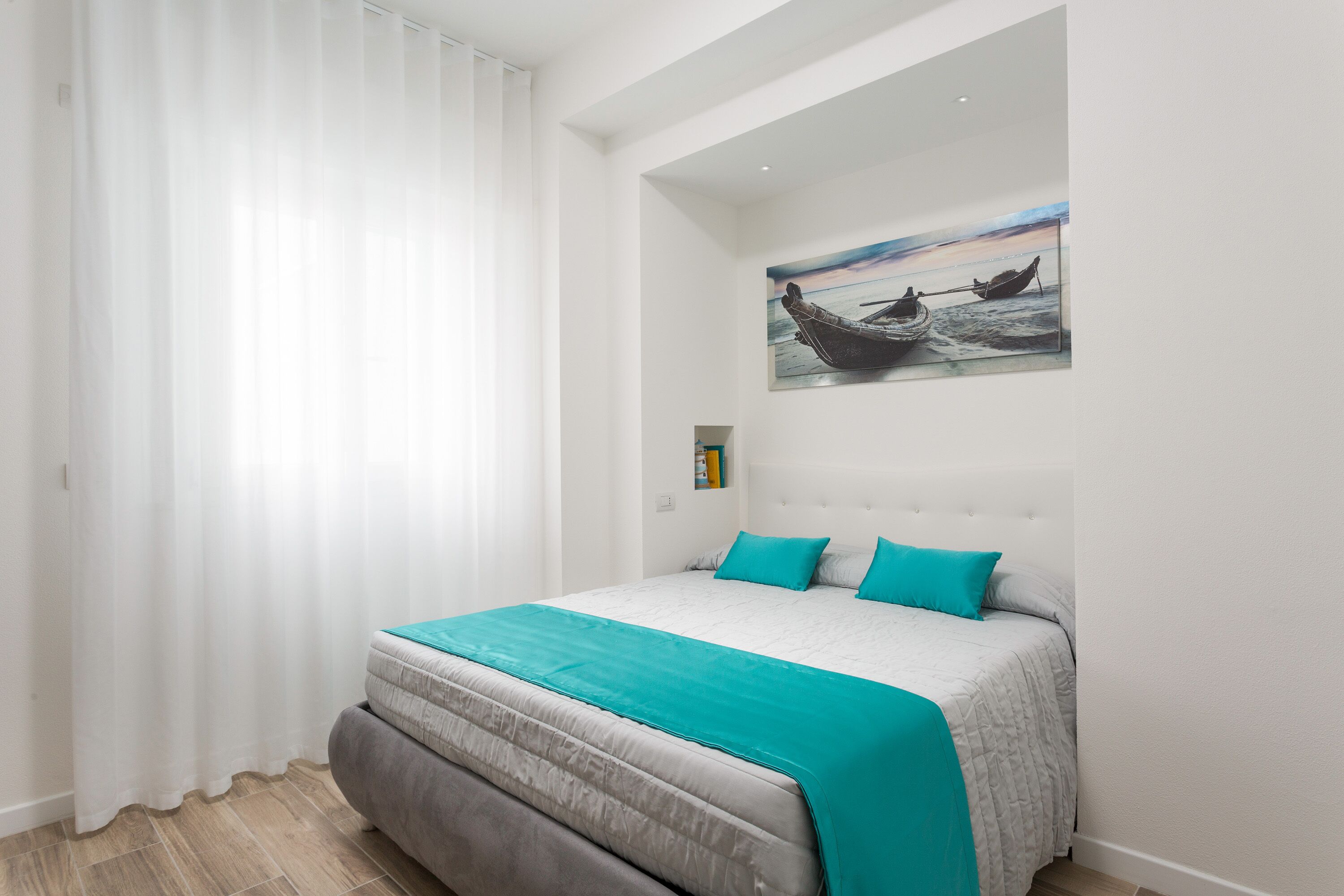 Ein Schlafzimmer mit einem großen Bett, weißen Wänden, einem Bootsbild an der Wand und einem Fenster mit weißen Vorhängen.