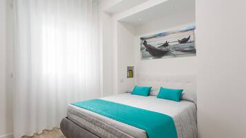 Ein Schlafzimmer mit einem großen Bett, weißen Wänden, einem Bootsbild an der Wand und einem Fenster mit weißen Vorhängen.