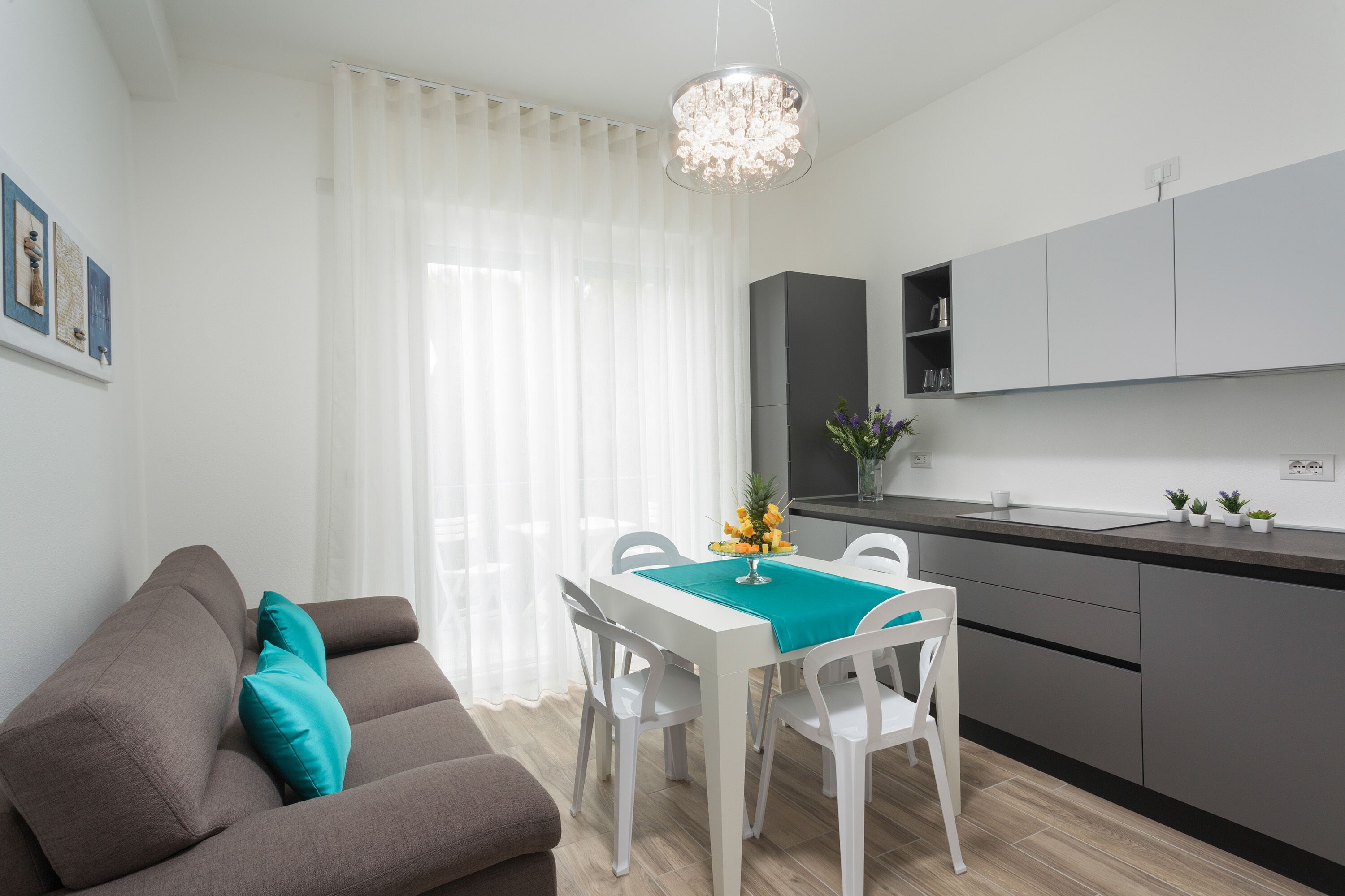 A modern kitchen with white cabinetry, a dining table with chairs, and a grey sofa with teal cushions.